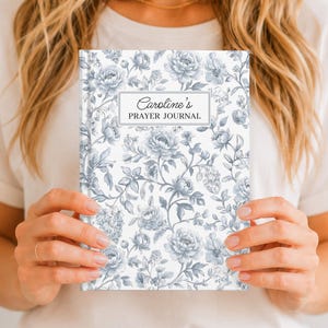 May include: A hardcover journal featuring a white background with a repeating pattern of blue-grey flowers and foliage. A rectangular label on the front cover contains the text Caroline's PRAYER JOURNAL in black lettering with a thin border.