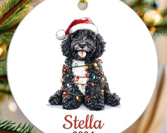 May include: A white ceramic Christmas ornament featuring a black and white dog wearing a red Santa hat and wrapped in colorful Christmas lights. The ornament is personalized with the name "Stella" and the year "2024".