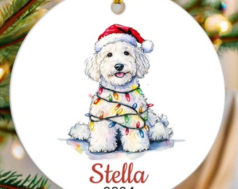 May include: A round Christmas ornament featuring a watercolor illustration of a white dog wearing a Santa hat and wrapped in colorful Christmas lights. The name "Stella" and the year "2024" are written in red script.