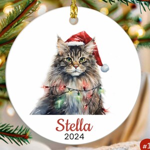 May include: A white ceramic Christmas ornament featuring a watercolor painting of a cat wearing a Santa hat and surrounded by Christmas lights. The cat's name, "Stella", and the year "2024" are printed below the image.