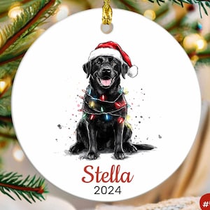 May include: A black Labrador Retriever wearing a red Santa hat and surrounded by colorful Christmas lights. The dog is sitting and the ornament has the name "Stella" and the year "2024" printed on it.