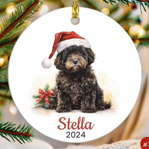 May include: A round white ceramic Christmas ornament featuring a brown and black dog wearing a red Santa hat. The dog is sitting with a red bow and green foliage behind it. The ornament is personalized with the name "Stella" and the year "2024".