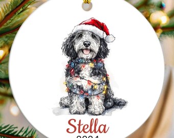 May include: A white ceramic Christmas ornament featuring a watercolor illustration of a dog wearing a Santa hat and string lights. The dog's name, "Stella," and the year "2024" are written in red script below the image. A gold string is attached for hanging.