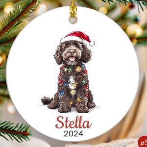 May include: A brown and white dog wearing a red Santa hat and wrapped in Christmas lights. The dog is sitting and looking at the camera. The ornament is personalized with the name "Stella" and the year "2024".