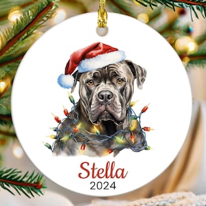 May include: A Christmas ornament featuring a gray dog wearing a red Santa hat and surrounded by colorful Christmas lights. The ornament is personalized with the name "Stella" and the year "2024".