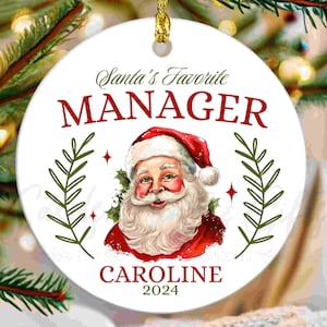 May include: White round Christmas ornament with a red and white illustration of Santa Claus. The ornament says "Santa's Favorite Manager Caroline 2024".