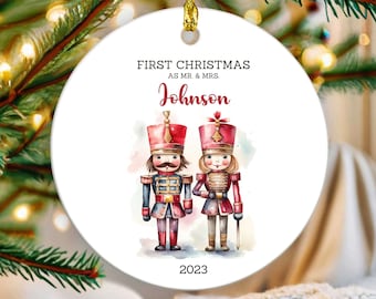 May include: A white, round Christmas ornament with a gold ribbon. The ornament features a watercolor illustration of a male and female nutcracker. Text reads "FIRST CHRISTMAS AS MR. & MRS. Johnson 2023."