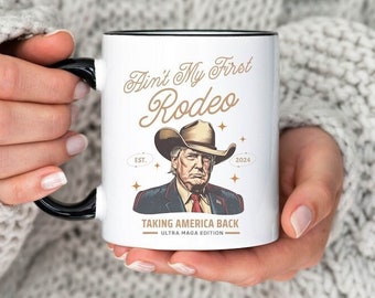 May include: White ceramic mug with a black handle. The mug features a cartoon illustration of Donald Trump wearing a cowboy hat and the text "Ain't My First Rodeo Est. 2024 Taking America Back Ultra MAGA Edition".