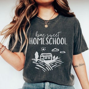 May include: A dark gray t-shirt with the text "home sweet HOMESCHOOL" in white. The shirt features a simple illustration of a house, sun, clouds, and trees in white.