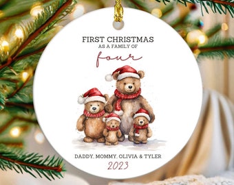 May include: A round Christmas ornament with a gold string. The ornament features a watercolor illustration of four teddy bears wearing Santa hats and scarves. Text reads "FIRST CHRISTMAS as a family of four" and names "Daddy, Mommy, Olivia & Tyler 2023."
