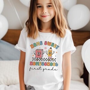 May include: A white t-shirt with a colorful graphic that says "Home Sweet Homeschool First Grade". The graphic features a cartoon book and pencil.