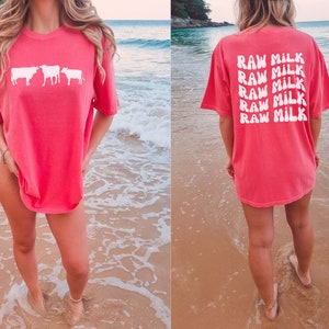 May include: A pink oversized t-shirt with a white graphic of three cows on the front and the text "RAW MILK" repeated five times on the back.