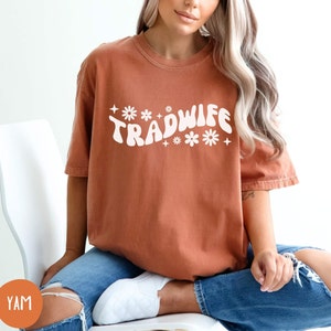 Puede incluir: Una camiseta de color &oacute;xido con el texto "TRADWIFE" en blanco con una fuente retro y detalles de flores blancas.