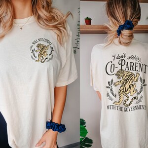 May include: A white t-shirt with a tiger graphic and the text "Will Not Comply" on the front and "I don't Co-Parent with the Government" on the back. The shirt is available in ivory.