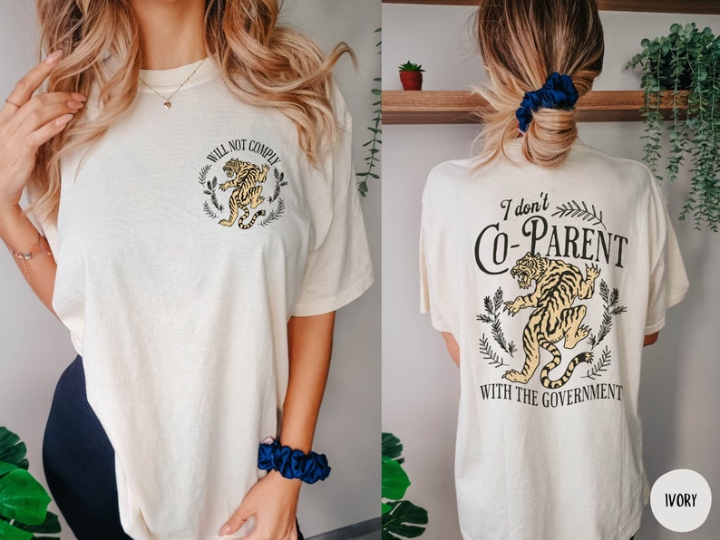 May include: A white t-shirt with a tiger graphic and the text "Will Not Comply" on the front and "I don't Co-Parent with the Government" on the back. The shirt is available in ivory.