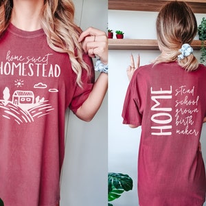 May include: A maroon t-shirt with the text "home sweet homestead" on the front and "home stead school grown birth maker" on the back.