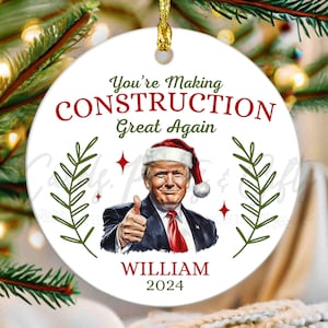May include: A white ceramic Christmas ornament with a gold string for hanging. The ornament features an image of a man wearing a Santa hat, with the text "You're Making CONSTRUCTION Great Again" and "WILLIAM 2024".