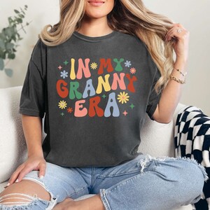 May include: A dark gray t-shirt with the text "In My Granny Era" in colorful, retro-style lettering. The text is surrounded by small, colorful flowers and stars.