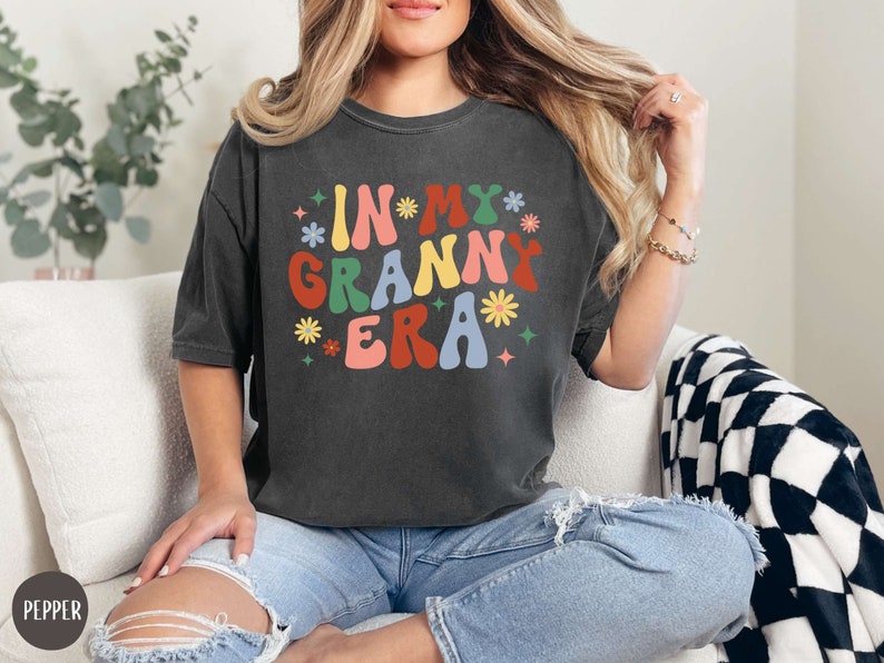 May include: A dark gray t-shirt with the text "In My Granny Era" in colorful, retro-style lettering. The text is surrounded by small, colorful flowers and stars.
