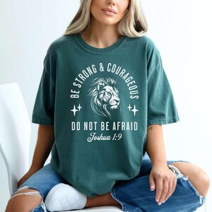 Pu&ograve; includere: Una maglietta blu turchese con una stampa grafica bianca di un leone con il testo "Be Strong & Courageous, Do Not Be Afraid, Joshua 1:9".