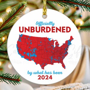 May include: A white round Christmas ornament with a red, white, and blue map of the United States. The text "Officially Unburdened by what has been 2024" is printed on the ornament.