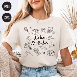 May include: Cream-colored t-shirt with the words "Wake & Bake" and baking-themed illustrations in black. The design includes a whisk, bread, bows, a bag of flour, a mixer, a rolling pin, an oven mitt, and hearts.