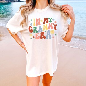 May include: A white t-shirt with the text "In My Granny Era" in colorful retro lettering with flowers and stars. The shirt is on a person standing on a beach.