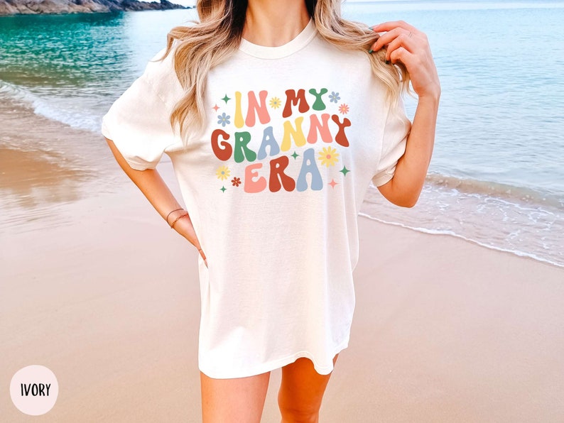 May include: A white t-shirt with the text "In My Granny Era" in colorful retro lettering with flowers and stars. The shirt is on a person standing on a beach.