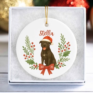 May include: White ceramic Christmas ornament featuring a black dog wearing a Santa hat, the name "Stella", and the year "2025". The ornament is decorated with a red bow, holly, and berries. The ornament is in a white gift box.