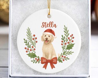 May include: White ceramic Christmas ornament featuring a light brown dog wearing a Santa hat, the name "Stella", and the year "2025". The dog is surrounded by a green and red wreath with a red bow at the bottom. The ornament is in a white gift box.