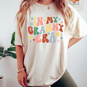 May include: A white t-shirt with the text "IN MY GRANNY ERA" in colorful retro-style lettering and flowers. The shirt is being worn by a person with long blonde hair and is paired with olive green biker shorts.