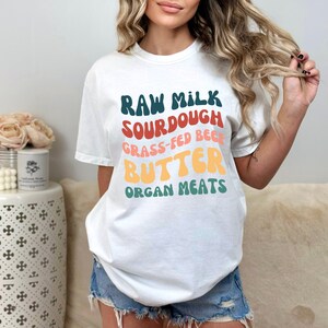 May include: White t-shirt with a colorful graphic design that reads "Raw Milk, Sourdough, Grass-Fed Beef, Butter, Organ Meats".