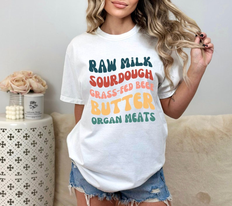 May include: White t-shirt with a colorful graphic design that reads "Raw Milk, Sourdough, Grass-Fed Beef, Butter, Organ Meats".
