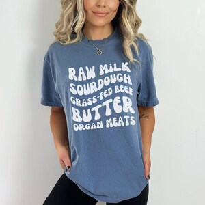 May include: A blue t-shirt with white text that reads "Raw Milk Sourdough Grass-Fed Beef Butter Organ Meats".