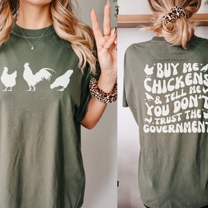 May include: Olive green t-shirt with a white graphic of three chickens on the front. The back of the shirt features the text "BUY ME CHICKENS & TELL ME YOU DON'T TRUST THE GOVERNMENT" in white, with chicken illustrations.