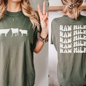 May include: A green t-shirt with a white silhouette of three cows on the front and the text "RAW MILK" repeated five times on the back.