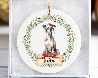 May include: A white ceramic Christmas ornament featuring a watercolor illustration of a greyhound wearing a Santa hat, surrounded by a green wreath with red berries. A bone-shaped tag with the name "Bella" is below the dog.