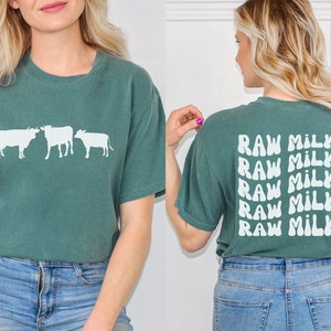 May include: A teal green t-shirt with a white silhouette of three cows on the front and the text "RAW MILK" repeated five times on the back.