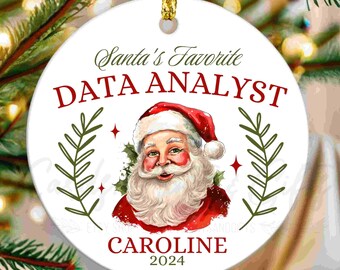 May include: A white Christmas ornament with a gold string features Santa Claus and the text "Santa's Favorite DATA ANALYST" in red and green. The name "CAROLINE" and the year "2024" are also included.