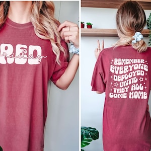 May include: A maroon t-shirt with white text. The front of the shirt says "RED friday" in a retro font. The back of the shirt says "REMEMBER EVERYONE DEPLOYED UNTIL THEY ALL COME HOME" in a retro font.