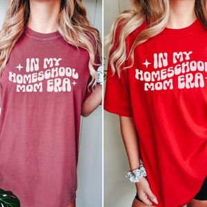 May include: Two women wearing oversized t-shirts with the text "In My Homeschool Mom Era" in a retro font. The shirt on the left is brick red and the shirt on the right is red.