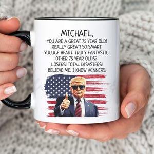 May include: White ceramic mug with a black handle and rim. The mug features a portrait of a man with sunglasses giving a thumbs up, set against an American flag design. Text includes "MICHAEL, YOU ARE A GREAT 75 YEAR OLD!" and other phrases.