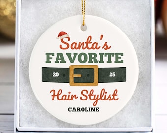 Puede incluir: Adorno navideño de cerámica blanca con el texto "Santa's Favorite Hair Stylist" en rojo y verde. El adorno presenta un gorro de Papá Noel y un diseño de cinturón con los años 20 y 25. El nombre "Caroline" está impreso en la parte inferior.
