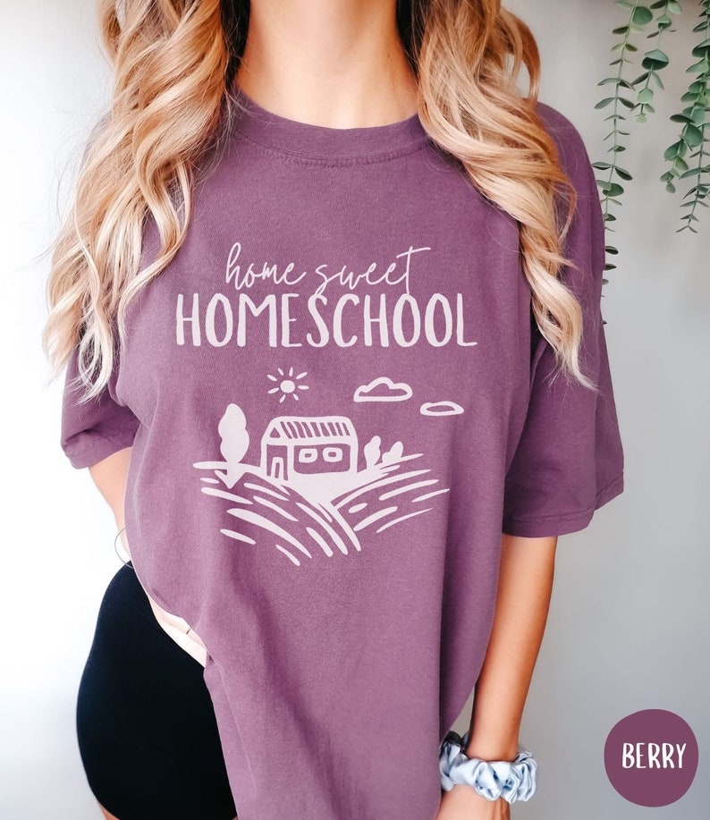 May include: A purple t-shirt with the text "home sweet HOMESCHOOL" in white. The shirt features a simple illustration of a house, sun, clouds, and trees.