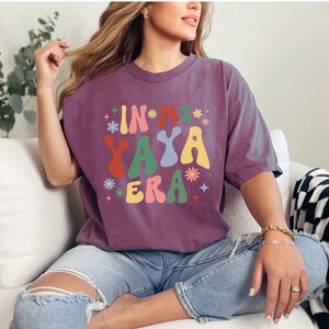 Puede incluir: Una camiseta morada con el texto "In My Yaya Era" en una fuente retro y colorida. La camiseta la lleva puesta una persona sentada en un sof&aacute; blanco.