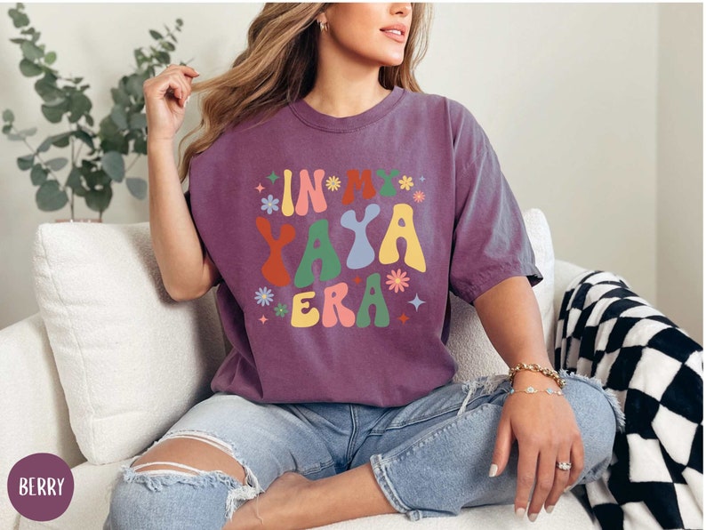 Puede incluir: Una camiseta morada con el texto "In My Yaya Era" en una fuente retro y colorida. La camiseta la lleva puesta una persona sentada en un sof&aacute; blanco.