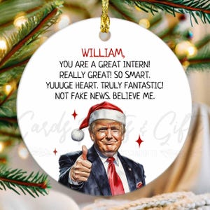 May include: A white, round Christmas ornament with a gold string for hanging. The ornament features a portrait of a man wearing a Santa hat and giving a thumbs-up. Text in red reads: "WILLIAM, YOU ARE A GREAT INTERN! REALLY GREAT! SO SMART. YUUUGE HEART. TRULY FANTASTIC! NOT FAKE NEWS. BELIEVE ME."