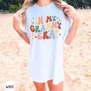 May include: White t-shirt with the text "In My Granny Era" in colorful retro font with flowers and stars.