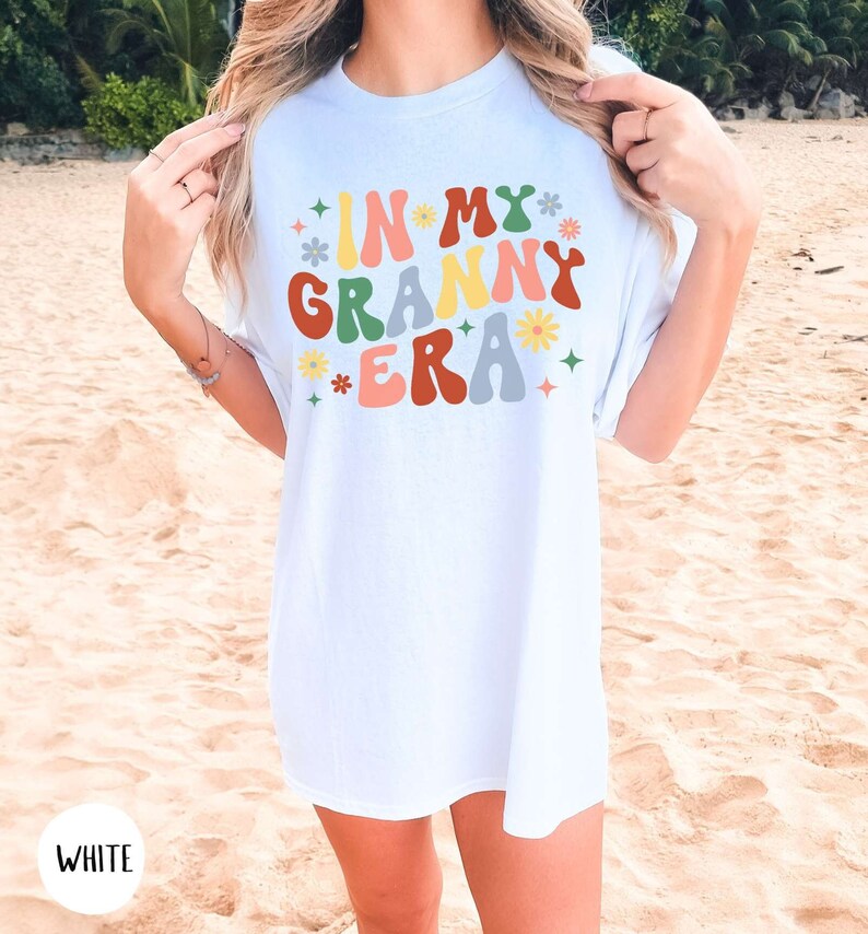 May include: White t-shirt with the text "In My Granny Era" in colorful retro font with flowers and stars.
