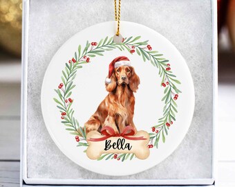May include: A round, white Christmas ornament featuring a brown dog wearing a Santa hat, centered within a green and red wreath. A bone-shaped tag with the name "Bella" is below the dog. The ornament is in a white gift box.